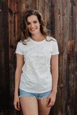 Junge Frau vor einer Holzwand stehend, die ein weißes T-Shirt trägt, auf dem das Logo von Kultsprache 